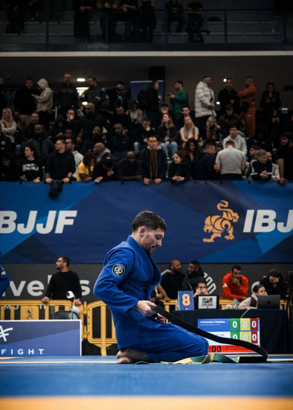 Compétiteur d'Akela Jiu Jitsu en kimono bleu nouant sa ceinture noire sur le tatami, devant les banderoles IBJJF et les tribunes