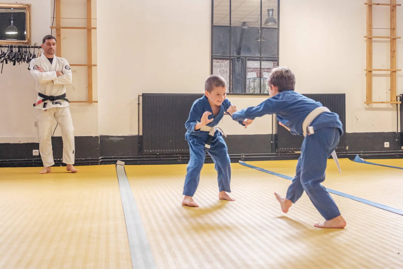 Deux enfants en kimono bleu s'affrontent au cours kids d'Akela Jiu Jitsu, sous l'oeil du professeur ceinture noire adossé au mur du dojo