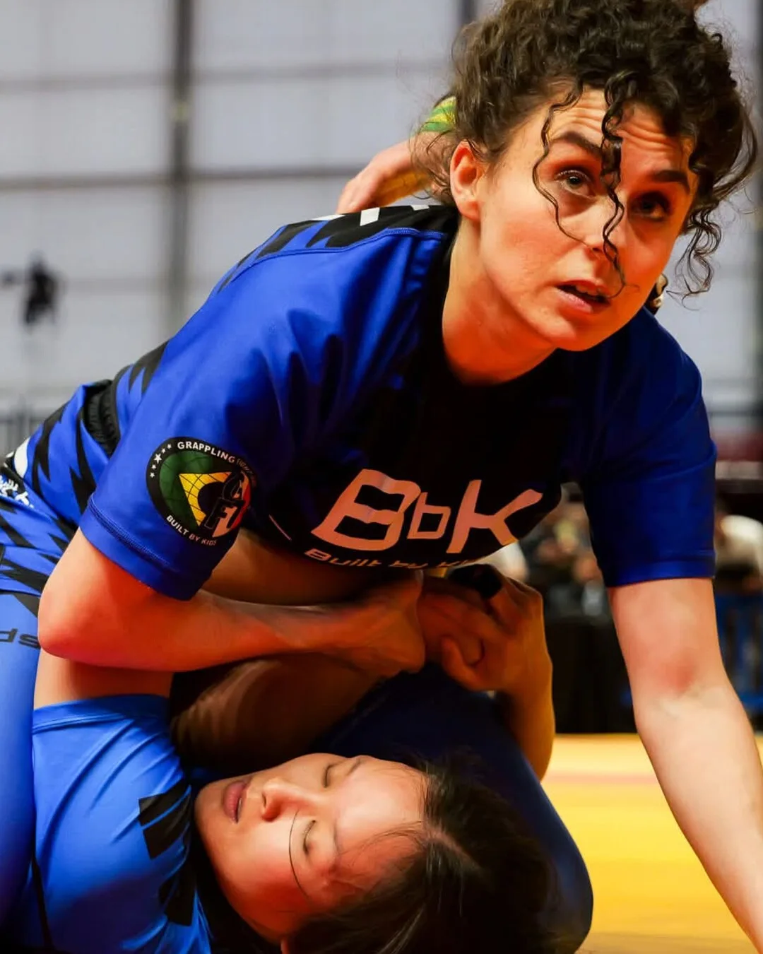 Compétitrice de Built by Kids en rashguard bleu floqué BbK, en contrôle sur son adversaire pendant un tournoi de grappling
