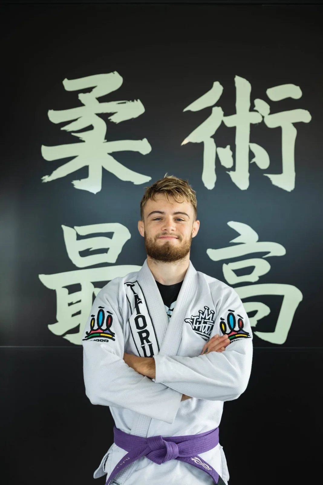 Clément Herrero, head coach JJB du Judo Jujitsu Colayrac, en kimono blanc et ceinture violette devant le mur de calligraphie japonaise du club