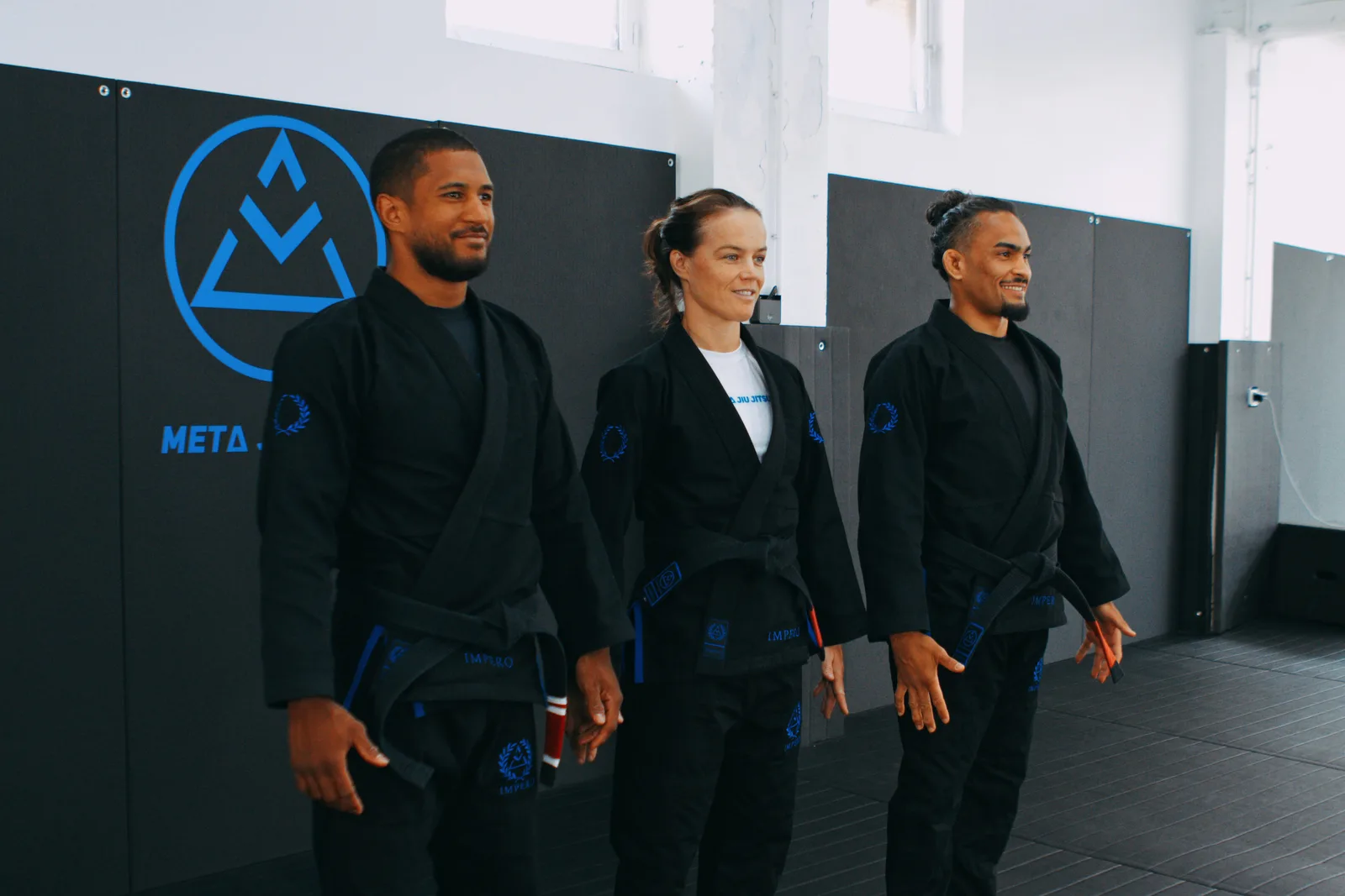 Les trois enseignants de Meta Jiu Jitsu, en kimono noir, alignés devant le mur au logo du club