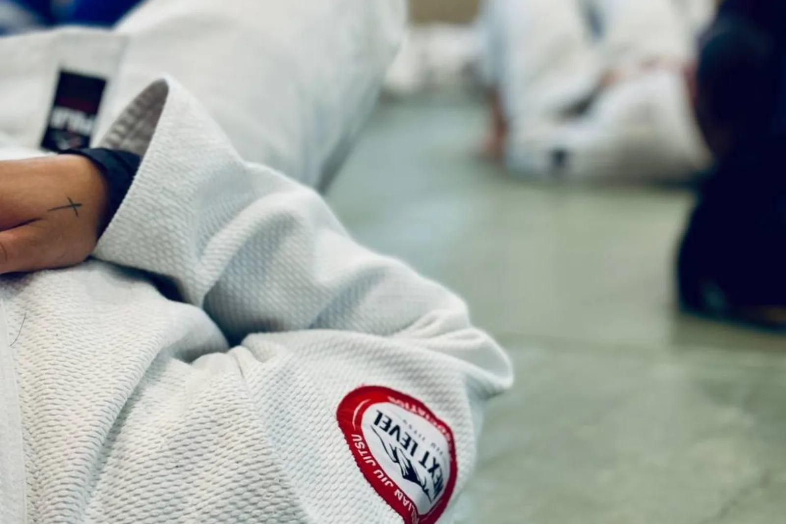 Gros plan sur le patch Next Level Jiu Jitsu cousu sur un kimono, des élèves à l'entraînement en arrière-plan flou