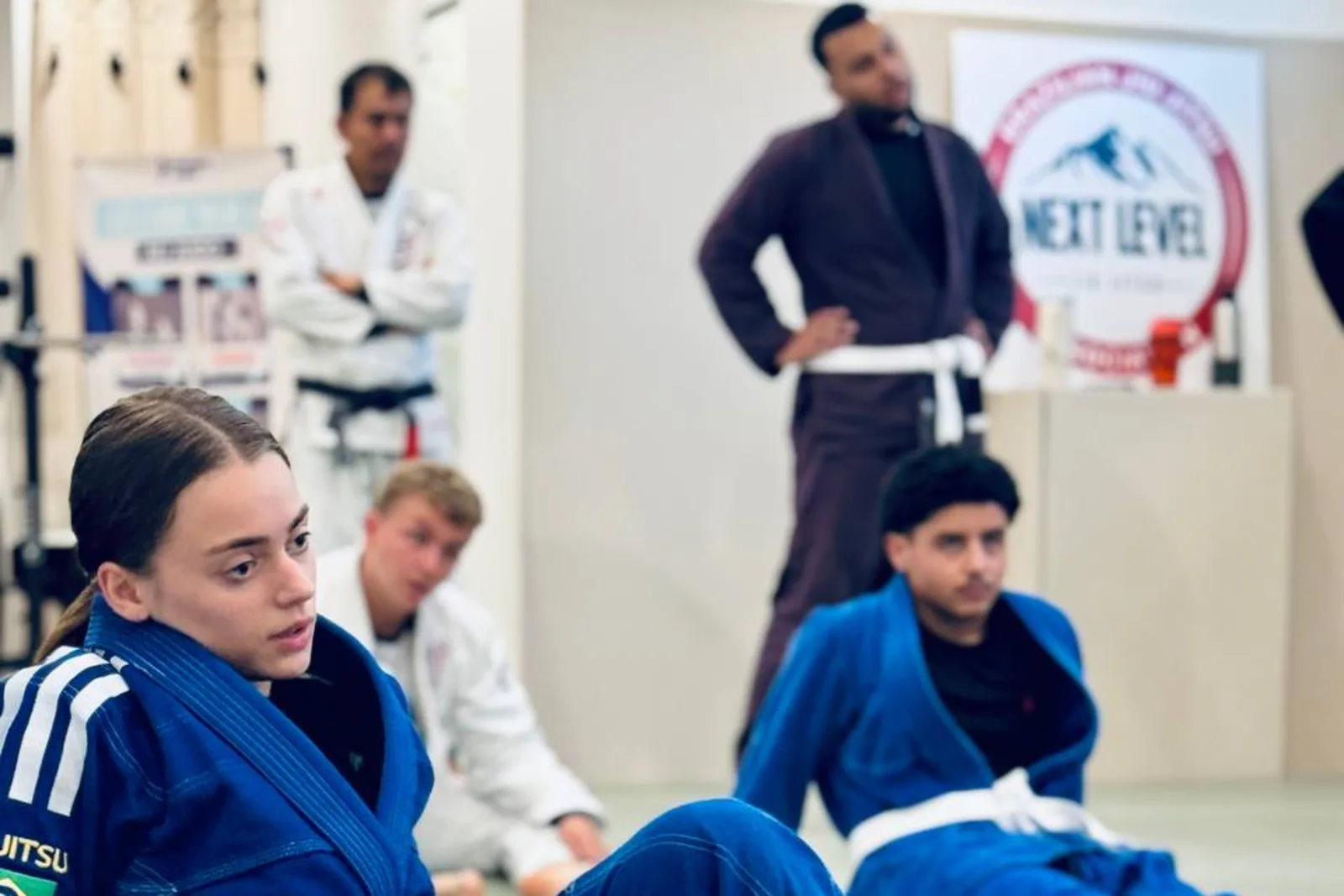 Des élèves de Next Level Jiu Jitsu Besançon attentifs pendant un cours, une élève en kimono bleu au premier plan et la bannière du club au fond