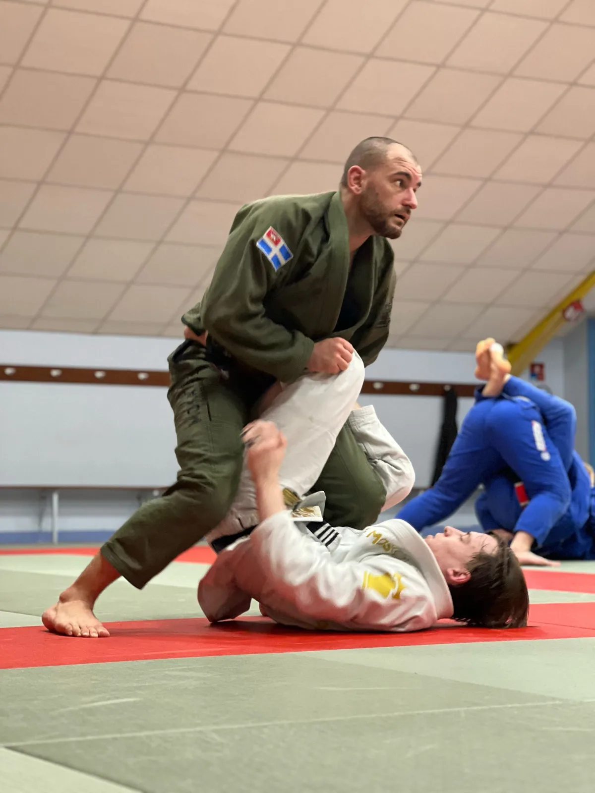 Sparring au sol pendant un cours du Saint-Malo Brazilian Jiu-Jitsu, passage de garde d'un pratiquant en gi kaki portant le drapeau malouin